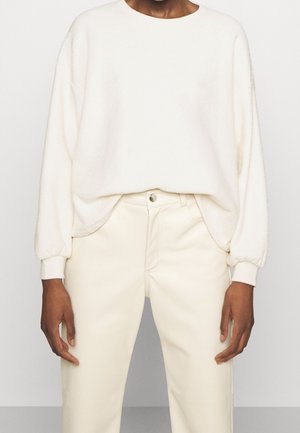 Person wearing a loose white long-sleeve sweater tucked slightly into high-waisted cream-colored pants, standing against a plain background.