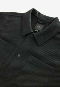 Černá prošívaná bunda s výrazným límcem, patentními druky a přední kapsou. Vyrobená z texturované látky s prošívaným vzorem.