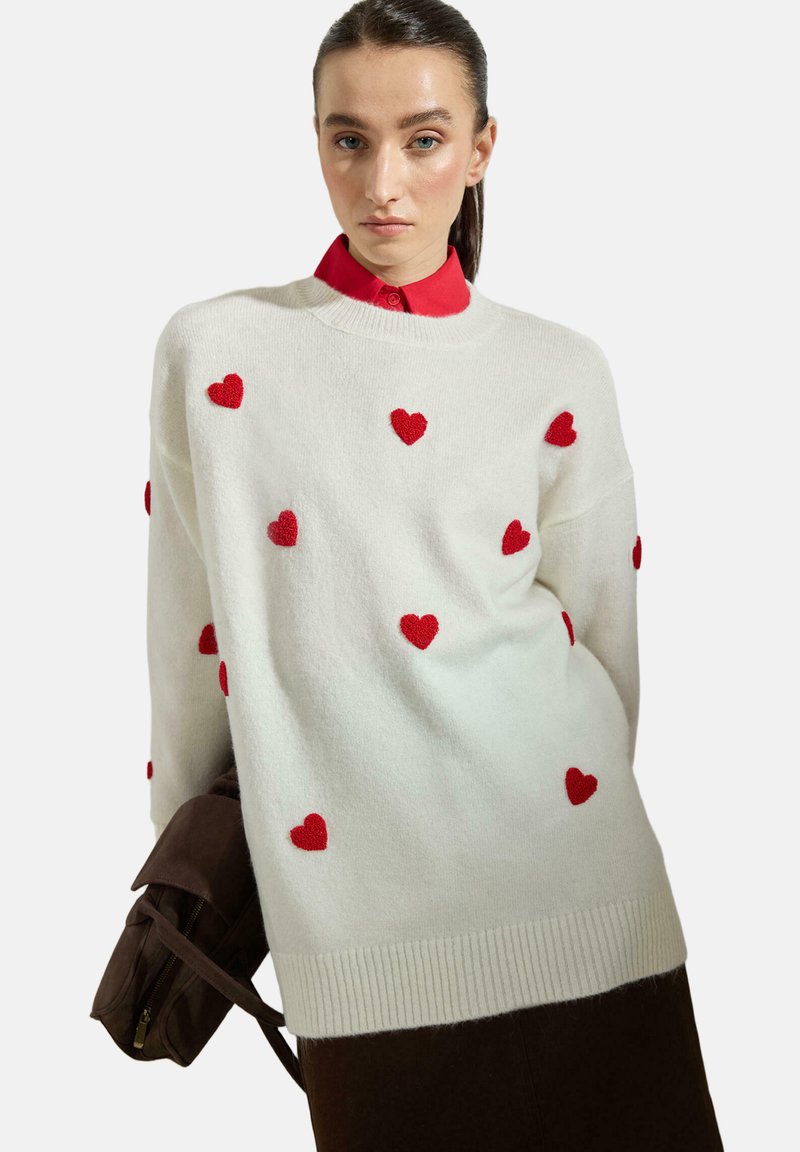 Femme portant un pull blanc orné de motifs de cœurs rouges sur une chemise rouge à col, tenant un sac marron devant un fond uni.