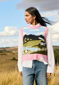 Eine Frau mit langen dunklen Haaren trägt eine Strickweste mit einem Landschaftsmuster in Rosa, Blau und Grün über einem weißen Shirt und Jeans und steht auf einem Feld.