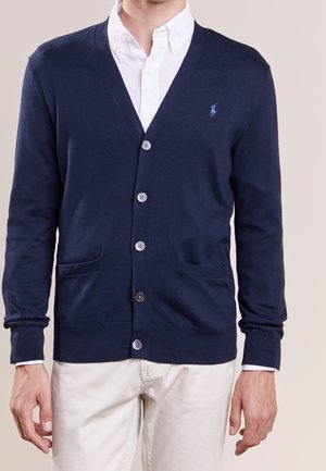 Mann trägt eine marineblaue, geknöpfte Strickjacke über einem weißen Hemd mit Kragen und hellbeiger Hose, steht vor einfachem Hintergrund.