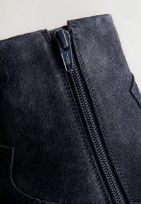 Stivale alla caviglia in suede nero con chiusura a zip, caratterizzato da dettagli di cucitura e una superficie liscia. La zip si estende sulla parte anteriore.
