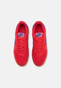 Paire de baskets rouges Nike avec logos Nike swoosh bleus, lacets rouges et semelles couleur gomme, vues de dessus sur un fond clair.