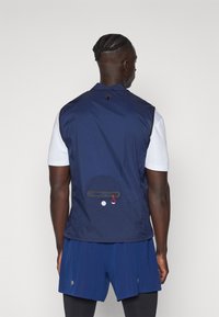 Gilet sans manches bleu marine fabriqué dans un tissu léger et texturé. Dispose d'une poche zippée à l'arrière et d'un passant pour l'attache.