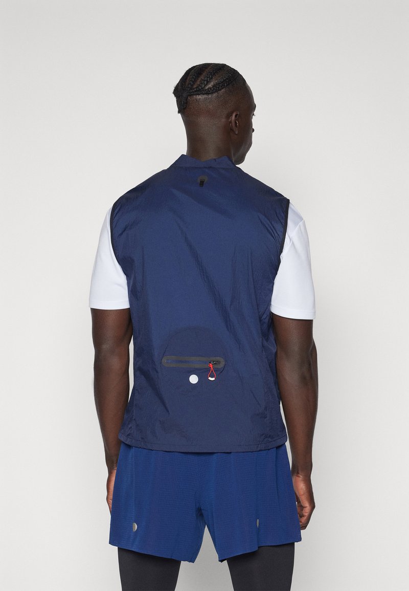 Gilet sans manches bleu marine fabriqué dans un tissu léger et texturé. Dispose d'une poche zippée à l'arrière et d'un passant pour l'attache.