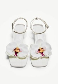 Vit sandal i syntetmaterial med en stor vit orkidédekoration med lila detaljer. Platt sula och justerbar vristrem.