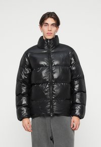 Jachetă puffer neagră cu finisaj lucios, guler înalt, fermoar frontal și secțiuni cu cusături orizontale. Purtați peste pantaloni de trening gri.