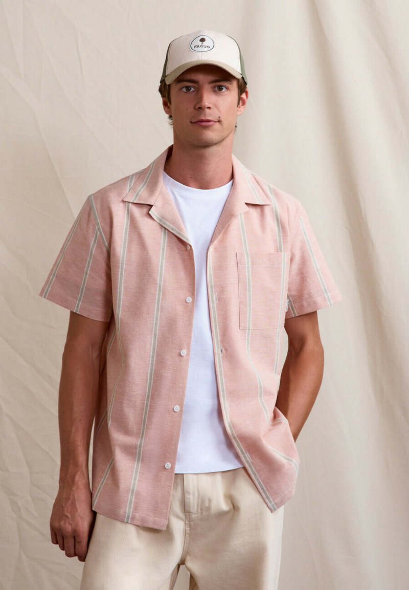 Jonge man staand, gekleed in een lichtroze gestreept kortärmlig shirt, wit T-shirt, beige broek en een beige pet met een logo.