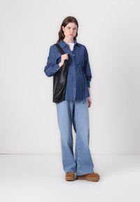 Camisa de botão de ganga em azul escuro, de corte largo, com um laço na cintura. Calças de ganga largas em azul claro combinadas com sapatos de plataforma castanhos e uma mala preta.