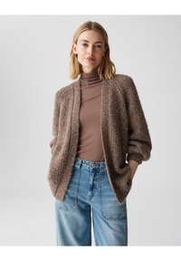 Cardigan marrone a pelo lungo con vestibilità morbida, apertura frontale e filato testurizzato. Indossato sopra un dolcevita marrone aderente e abbinato a jeans a vita alta.