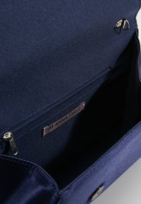 Sac à main en tissu bleu marine à texture lisse, avec une poche zippée à l'intérieur et une étiquette en métal. Fermeture à boucle ronde.