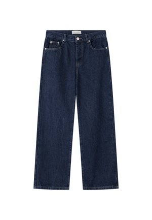 Ung mand med kort blond hår iført en hvid t-shirt, mørke løse jeans og hvide sneakers, der kigger ned, mens han holder i sin skjortekant.