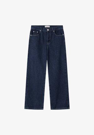 Jonge man met kort blond haar, die een wit T-shirt, donkere losse jeans en witte sneakers draagt, kijkt naar beneden terwijl hij de zoom van zijn T-shirt vasthoudt.