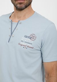 Lyseblå kortermet skjorte med henley-hals med tre sølvfargede trykknapper og trykt tekst i mørkeblå og røde detaljer.