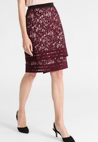 Burgunderfärgad spetskjol i pennform med lagerdesign och elastisk midja; genomskinligt blommönster; knä-lång, matchad med svarta klackar.