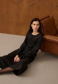 Femme aux cheveux foncés portant une longue robe noire, assise sur le sol, adossée à un fauteuil en cuir marron, fond neutre.