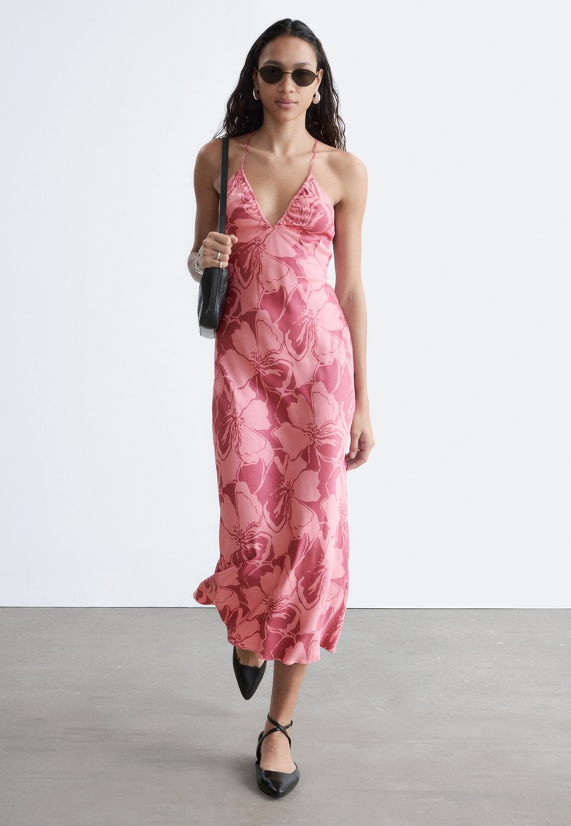Femme portant une robe nuisette fleurie rose, des chaussures pointues noires, un sac noir, des lunettes de soleil rondes et des boucles d’oreilles anneaux marchant sur un sol gris.