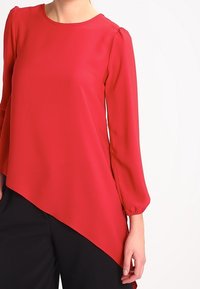 Blusa roja de manga larga con cuello redondo y dobladillo asimétrico. Fabricada en tela suave con un ligero volumen en los hombros.