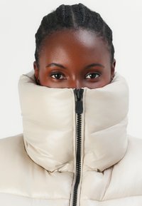 Beige pufferjack met een hoge kraag, voorzien van een zwarte rits en een geborduurd textuur die het gezicht van de drager bedekt. Glad, glanzend materiaal.
