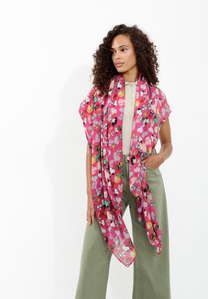 Écharpe rose avec des motifs de personnage de dessin animé, en tissu léger et transparent avec un tombé fluide, portée sur un haut beige et un pantalon vert.