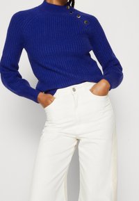 Pull en tricot bleu vibrant avec un col montant, un design côtelé texturé, et trois boutons noirs décoratifs sur l'épaule. Associé à un pantalon large blanc.