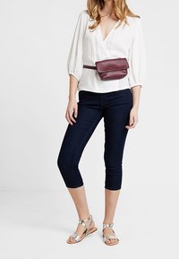 Mujer con blusa blanca, jeans oscuros cortos, sandalias plateadas y un bolso de cuero burdeos en la cintura.