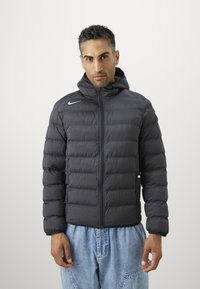 Sort puffer-jacket med lynlås foran, hætte og vandrette baffle. Har et hvidt logo på venstre bryst og elastiske ærmekanter.