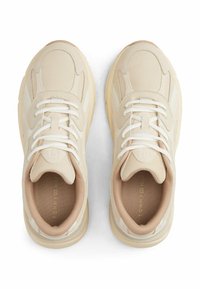Baskets beiges avec des panneaux lisses et texturés, au design incurvé, lacets blancs et doublure douce. Présente un logo et un detailing minimaliste.