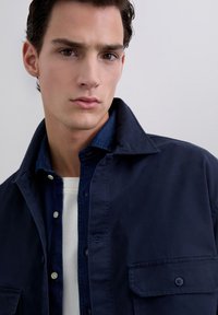 Un joven con cabello oscuro viste una chaqueta navy de capas sobre una camisa de mezclilla y una camiseta blanca, mirando ligeramente hacia el lado contra un fondo blanco liso.