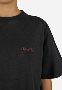 Svart bomull t-skjorte med kort erm, med rødt brodert tekst "Values & Visions" på venstre brystområde. Enkelt og uformelt design.