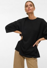 Ženska z zavezanimi lasmi, oblečena v široko črno kratko majico, rjave hlače, dolge uhane in zapestnice, ki pozira z rokami na boku.