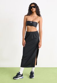 Top bandeau preto com o logótipo branco da Adidas, combinado com uma saia preta até ao tornozelo com uma fenda lateral. Sapatos desportivos escuros com detalhes brancos.