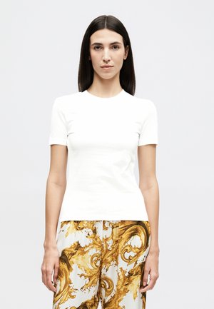 Νεαρή γυναίκα με ίσια σκούρα μαλλιά, φοράει απλό λευκό κοντομάνικο πουκάμισο και λευκό παντελόνι με περίτεχνο χρυσό μπαρόκ σχέδιο.