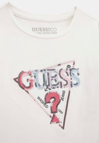 Bílý tričko z organické bavlny s texturovaným, flitrovým trojúhelníkovým logem se „GUESS“ v šedé, růžové a červené a textem „WASHED JEANS 1203“. 