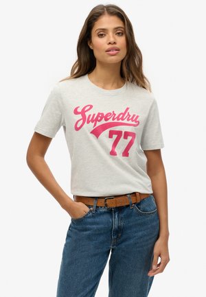 T-shirt en coton gris clair avec le grand texte rose "Superdry 77", manches courtes et col rond, porté avec un jean bleu et une ceinture marron.