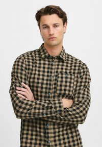 Cămașă cu carouri maro și negre, din flanel, cu un guler cu nasturi, un singur buzunar pe piept și nasturi albaștri.