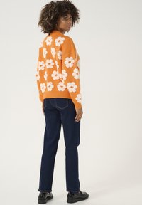 Narancssárga kötött pulóver, amely nagy fehér virágmintákkal rendelkezik, sötétkék egyenes szárú farmernadrággal és fekete cipővel párosítva, hátulról nézve.