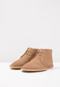 Stivaletti in suede marrone alla caviglia con punta appuntita, design con lacci e dettagli cuciti attorno alla suola. Texture liscia e silhouette minimalista.