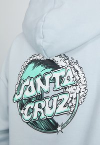 Sweatshirt azul claro com um grande gráfico em preto e teal de uma onda, acompanhado do texto "Janta Cruz" em uma fonte ousada e divertida.