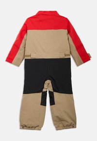Kinderen jumpsuit met een rode bovenkant, beige middenstuk en zwarte onderkant. Gemaakt van een gladde stof met elastische boorden en klittenbandaccenten.