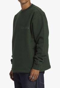 Grön sweatshirt med rund hals, ribbade muddar och en synlig logga i en mörkare grön nyans. Har en sidoficka och en slät textur.