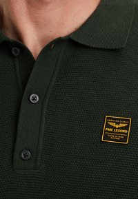 Dunkelgrünes Polohemd aus strukturiertem Stoff, zwei schwarze Knöpfe und ein gelbes Markenpatch mit der Aufschrift "PME Legend."