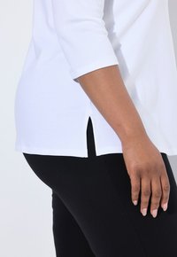 Weißes Oberteil mit drei Viertelärmeln und seitlichen Schlitzen am Saum, glatte Textur, kombiniert mit schwarzen Leggings, die eine figurbetonte Silhouette zeigen.
