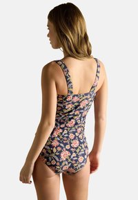 Žena otočená chrbtom, oblečená v námornícky kvetinovom jednodielnom plavkách s širokými ramienkami na pozadí svetlej farby.