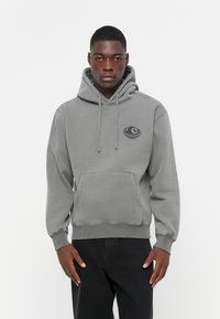 Grijze hoodie met een voorzak, trekkoordkap en een ronde logo patch op de borst. Gemaakt van zachte stof met ribbelmanchetten en onderrand.