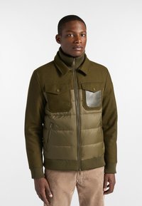 Giacca verde oliva con colletto a maglia e parte inferiore imbottita. Presenta due tasche sul petto, una chiusura con zip e polsini a costine.
