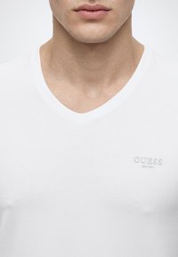 Κοντινό πλάνο ενός άντρα που φοράει λευκό T-shirt με V-λαιμόκοψη και τη λέξη "GUESS" κεντημένη στο αριστερό μέρος του στήθους.