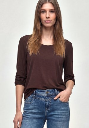 Mujer con cabello largo castaño claro, vestida con una camisa marrón de manga larga y jeans azules, de pie con una mano en el bolsillo frente a un fondo liso.