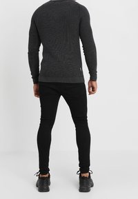 Dunkelgrauer, gerippter Strickpullover mit langen Ärmeln, kombiniert mit schwarzen Slim-Fit-Hosen und schwarzen Sportschuhen, von hinten gesehen.