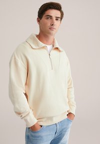 Kreminės spalvos zip-up džemperis, pagamintas iš minkštos medžiagos, turi plačią apykaklę ir laisvą kirpimą. Dėvimas ant šviesiai mėlynų džinsų.